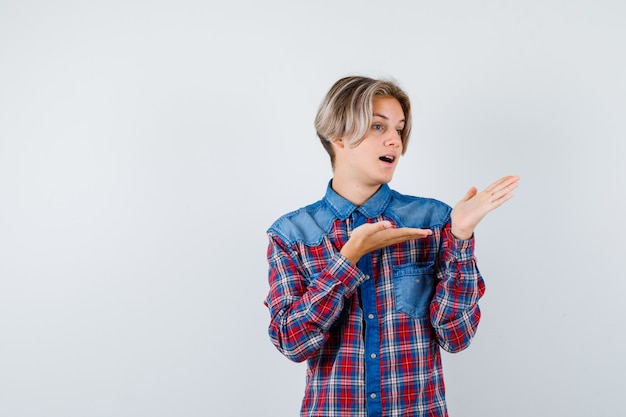 Teen boy pretending to show something in checkered shirt and looking perplexed , front view.