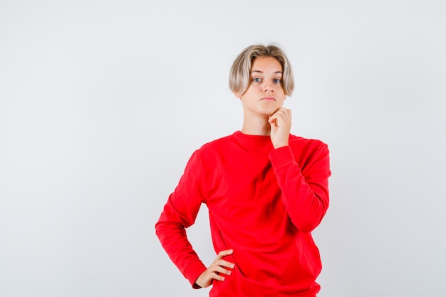 Teen boy in red sweater keeping finger on chin and looking thoughtful , front view.