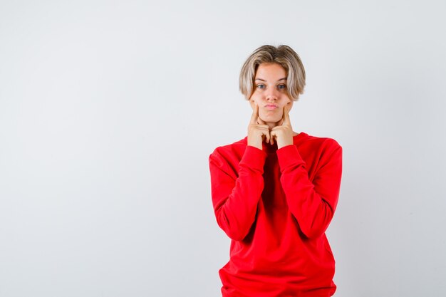 Teen boy in red sweater keeping fingers on cheeks, pouting lips and looking comic , front view.