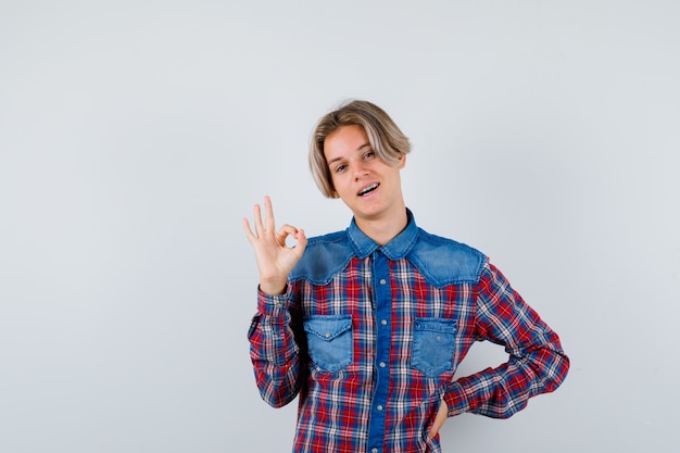 Teen boy showing ok sign in checkered shirt and looking joyful , front view.