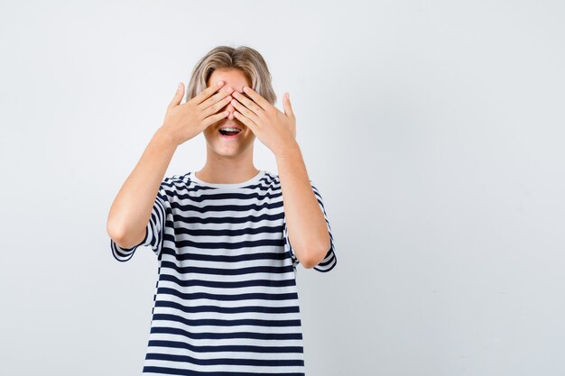 Teen boy in t-shirt covering eyes with hands, opening mouth and looking excited , front view.