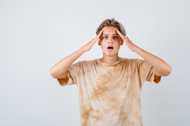 Teen boy with hands on forehead, opening mouth in t-shirt and looking terrified , front view.