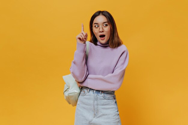 Teen cool brunette Asian woman in eyeglasses purple sweater and denim skirt points up has great new idea and poses with backpack on orange background