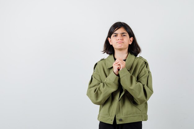 アーミーグリーンのジャケットを着た10代の少女は、祈りのジェスチャーで手を握りしめ、暗い、正面図を探しています。