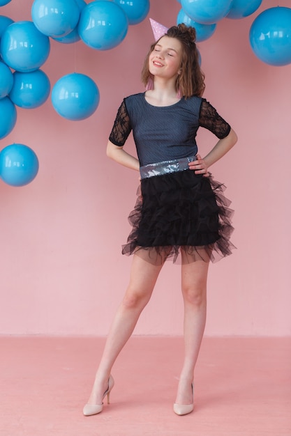 Teen girl in birthday cone posing on pink wall and blue balloons backdrop.