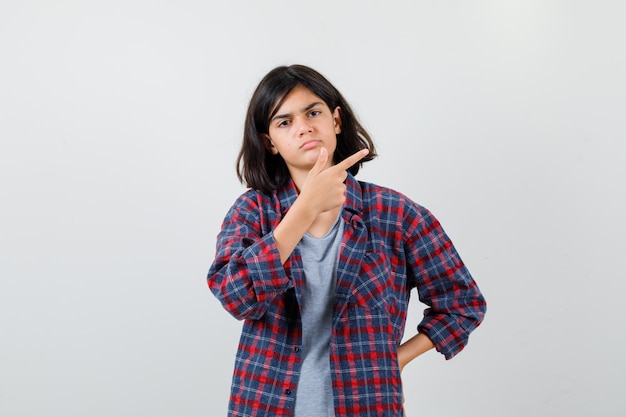 Ragazza teenager in abiti casual che punta all'angolo in alto a destra e sembra cupa, vista frontale.