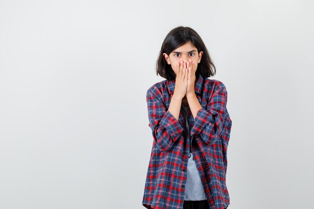 Teen girl in checkered shirt covering mouth with hands and looking scared , front view.