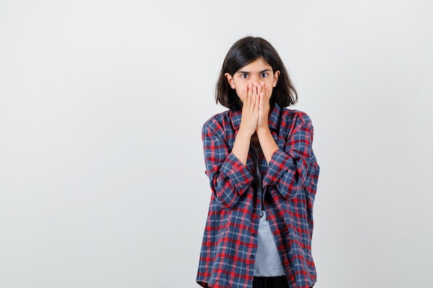 Teen girl in checkered shirt covering mouth with hands and looking scared , front view.