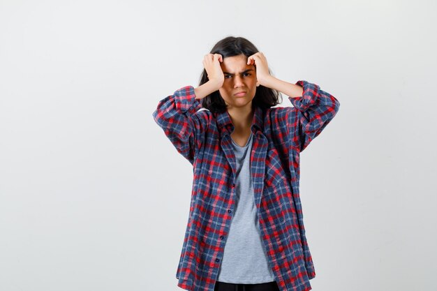 Teen girl in checkered shirt holding hands near face and looking morose , front view.