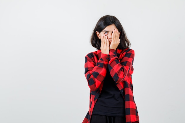 Teen girl covering eye with hand while pulling down her skin in t-shirt, checkered shirt and looking bored. front view.