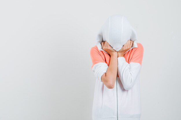 Teen girl covering her head and face with cap in white jacket and looking hidden.