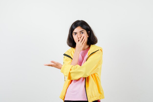 Teen girl covering mouth with hand, spreading arm in yellow tracksuit, t-shirt and looking surprised , front view.