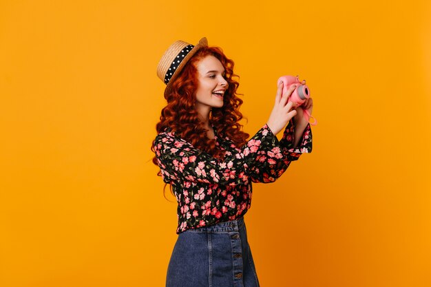 Teen girl in denim skirt and stylish blouse happily makes photo, holding mini camera on orange space.
