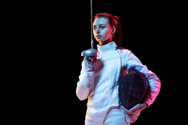 Teen girl in fencing costume with sword in hand isolated on black background, neon light.