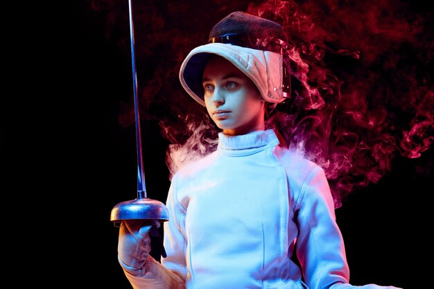 Teen girl in fencing costume with sword in hand isolated on black wall
