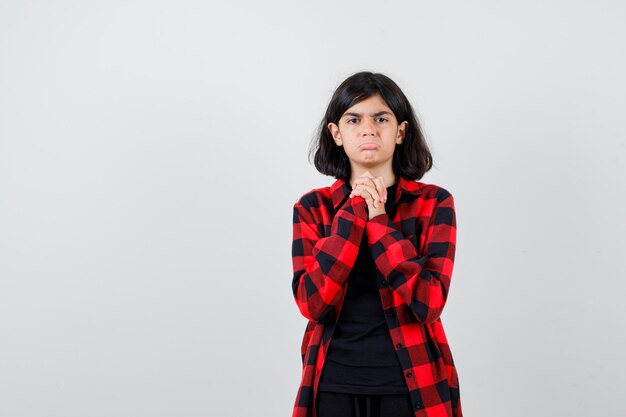 Teen girl holding clasped hands in pleading gesture, curving lower lip in casual shirt and looking grumpy , front view.