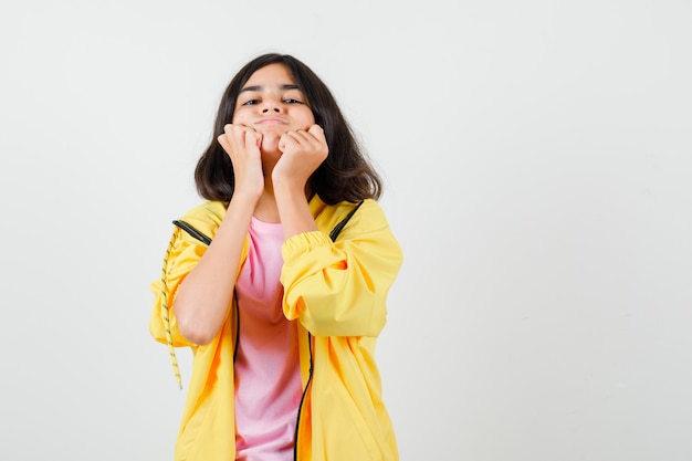 Ragazza teenager che tiene i pugni sulle guance in tuta gialla, t-shirt e sembra dispiaciuta, vista frontale.