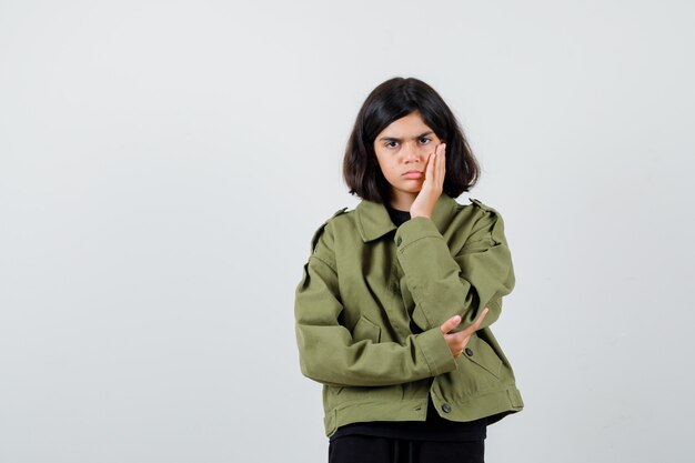 Teen girl holding hand on cheek in t-shirt, jacket and looking wistful. front view.