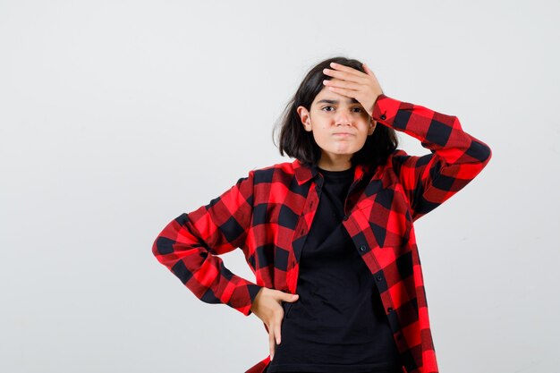 Teen girl holding hand on forehead in t-shirt, checkered shirt and looking forgetful , front view.