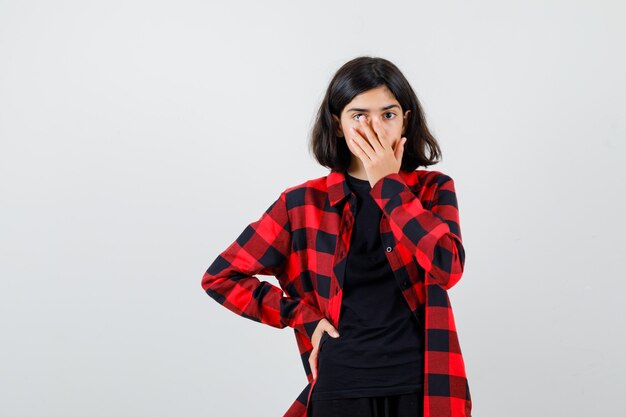 Teen girl holding hand on mouth in t-shirt, checkered shirt and looking puzzled. front view.