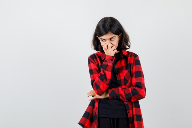 Teen girl holding hand on mouth in t-shirt, checkered shirt and looking thoughtful. front view.