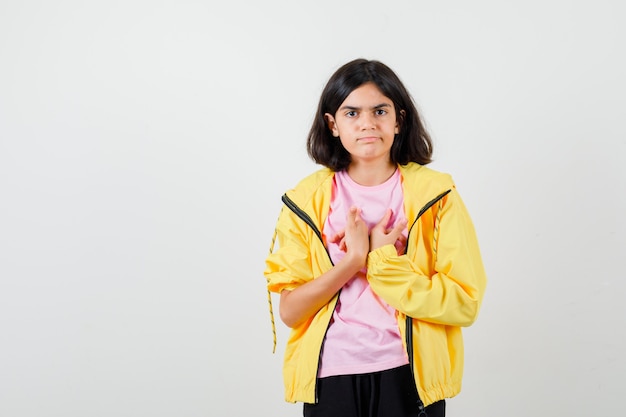 Ragazza teenager che si tiene per mano sul petto in t-shirt, giacca e sembra premurosa. vista frontale.