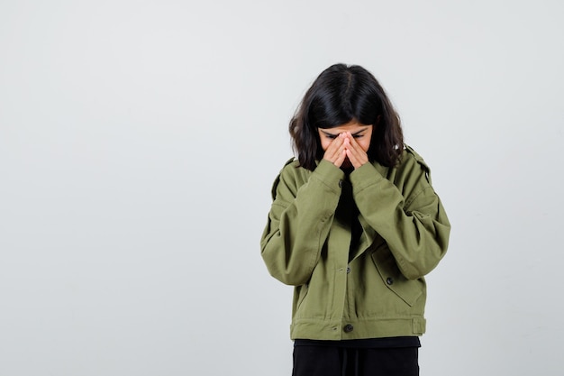 Free photo teen girl holding hands on face in army green jacket and looking mournful , front view.