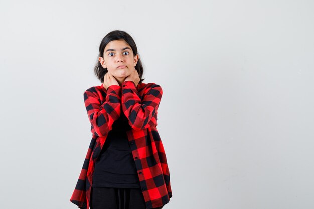 Teen girl holding hands on neck in t-shirt, checkered shirt and looking scared. front view.