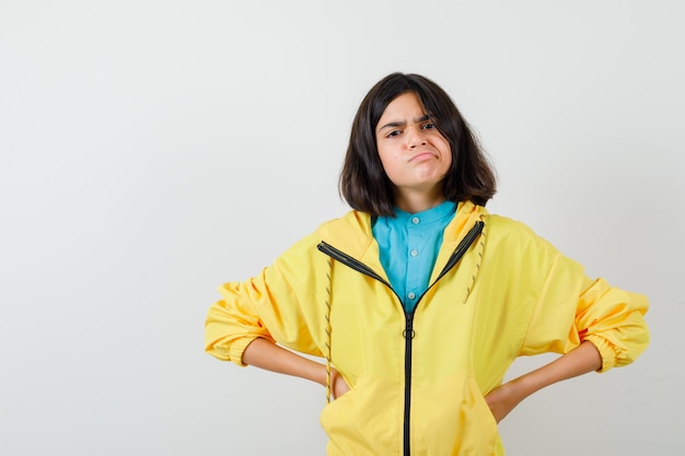 Free photo teen girl holding hands on waist while frowning in yellow jacket and looking confused , front view.