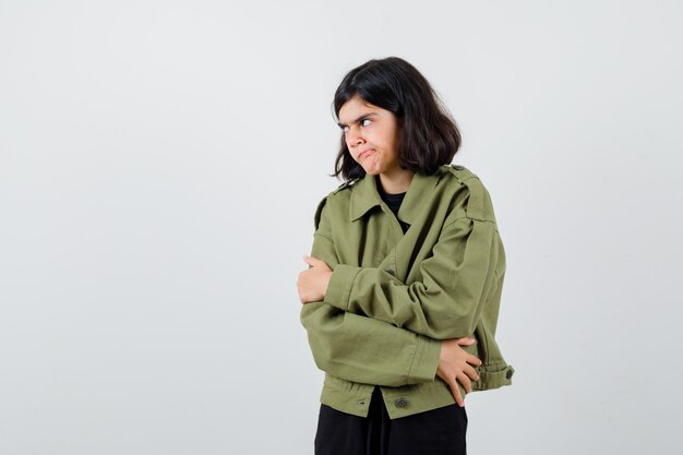 Teen girl hugging herself, looking aside, curving lips, frowning face in army green jacket and looking spiteful , front view.