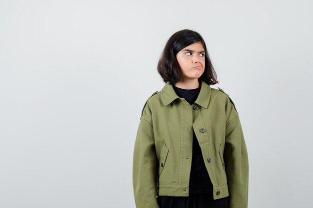 Teen girl looking aside while curving lips in army green jacket and looking displeased , front view.