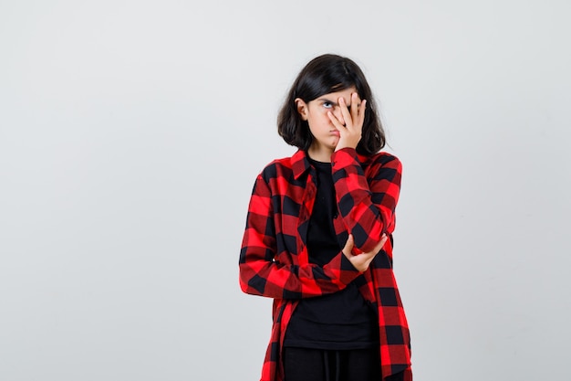 Teen girl looking through fingers in casual shirt and looking bored , front view.