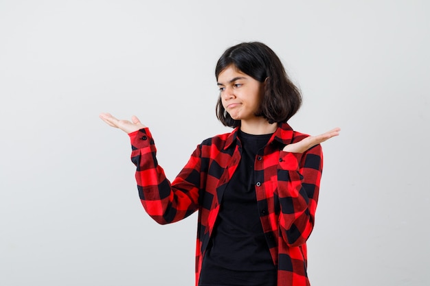 Teen girl making scales gesture in t-shirt, checkered shirt and looking pensive , front view.