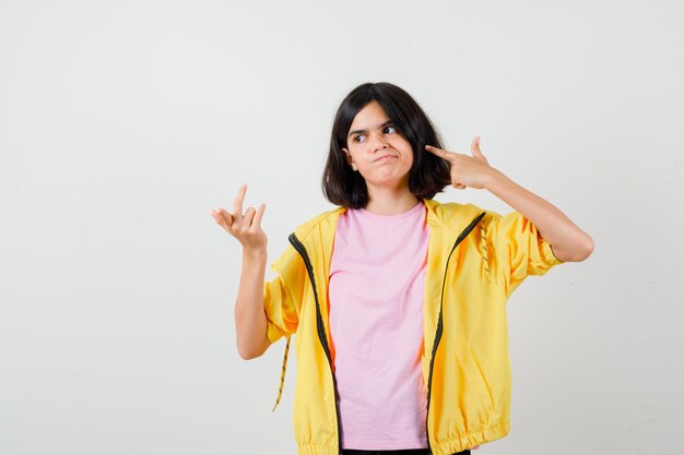 Teen girl making suicide gesture, raising hand in puzzled gesture in t-shirt, jacket and looking hesitant , front view.