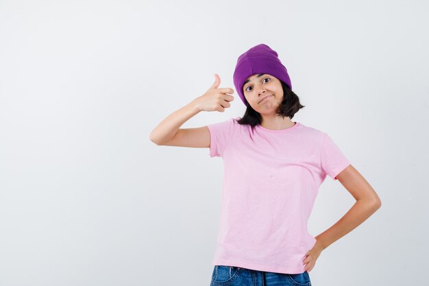 Teen girl in pink t-shirt, beanie, jeans showing thumb up, holding hand on waist and looking cheerful , front view.