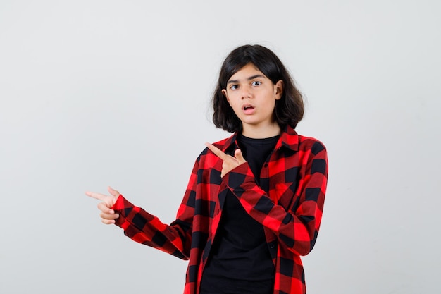 Teen girl pointing aside in t-shirt, checkered shirt and looking perplexed. front view.