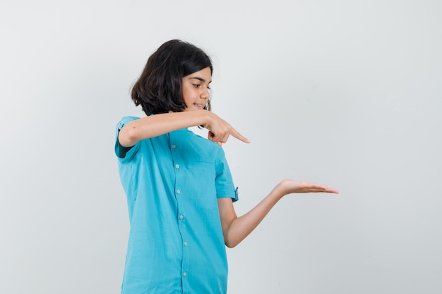 Teen girl pointing at her spreading palm in blue shirt and looking focused.