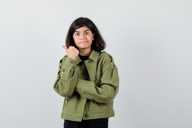 Tシャツ、緑のジャケットの親指で左側を指して、注意深く見ている10代の少女。正面図。