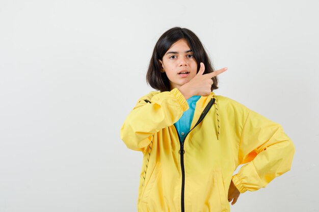 Teen girl pointing right while keeping hand on waist in shirt, yellow jacket and looking cheerless. front view.