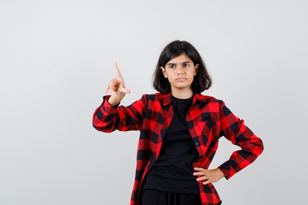 Teen girl pointing up in casual shirt and looking pensive. front view.