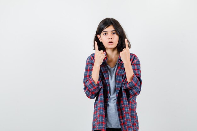 Teen girl pointing up in checkered shirt and looking surprised , front view.