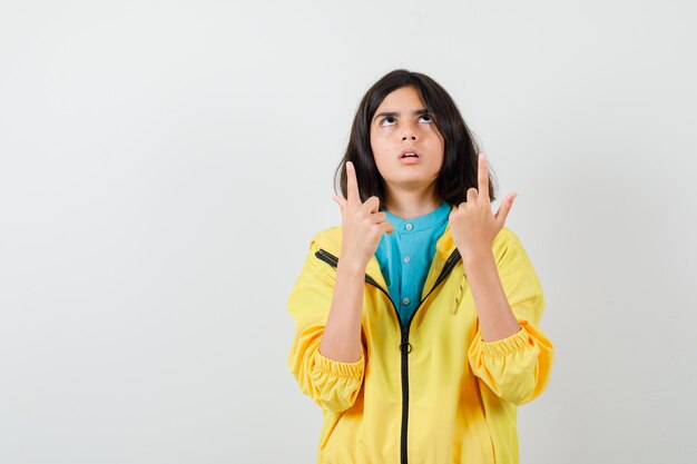 Teen girl pointing up, looking upward in yellow jacket and looking puzzled. front view.