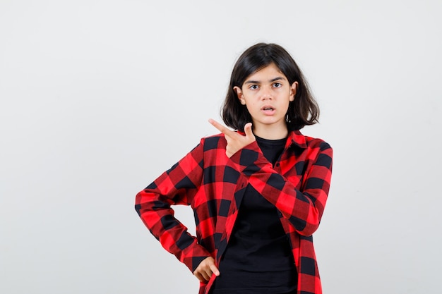 Teen girl pointing at upper left corner in t-shirt, checkered shirt and looking perplexed , front view.