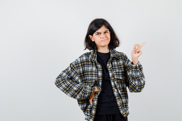 Teen girl pointing at upper right corner, holding hand on waist in casual shirt and looking disappointed. front view.