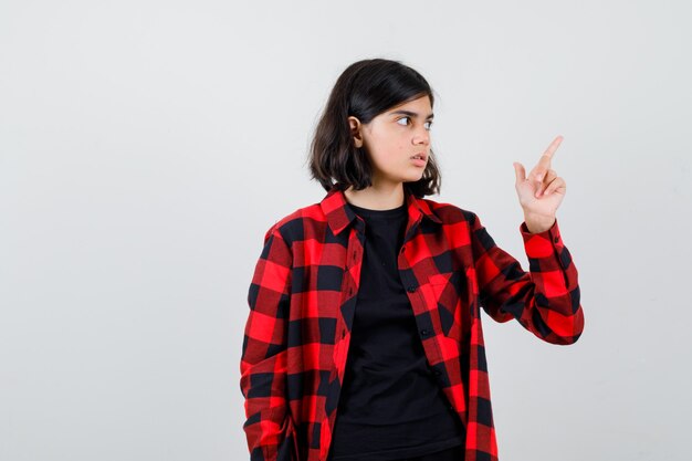 Teen girl pointing at upper right corner in t-shirt, checkered shirt and looking perplexed. front view.