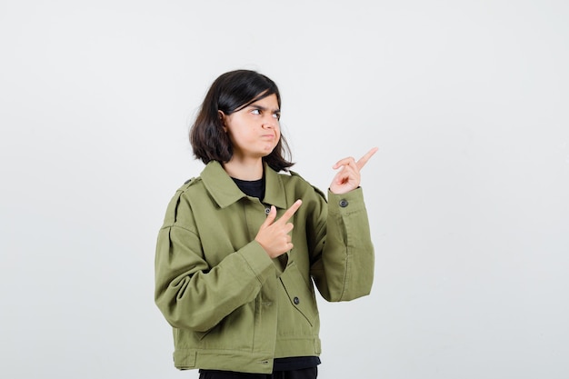 Teen girl pointing at upper right corner in t-shirt, jacket and looking indecisive , front view.