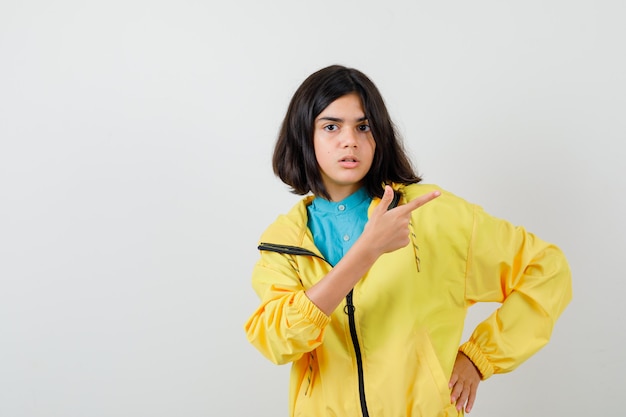 Free photo teen girl pointing at upper right corner in yellow jacket and looking puzzled. front view.