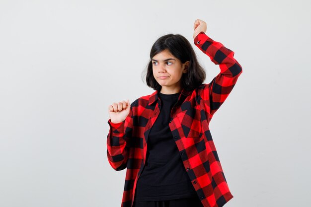 Teen girl raising fists spread in t-shirt, checkered shirt and looking dissatisfied , front view.
