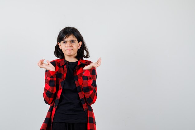 Teen girl raising hands aside in t-shirt, checkered shirt and looking disappointed , front view.