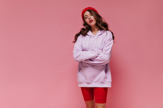 Teen girl in red hat and cycling shorts poses on pink background Portrait of curly attractive woman with bright lips dressed in purple oversized hoodie on isolated